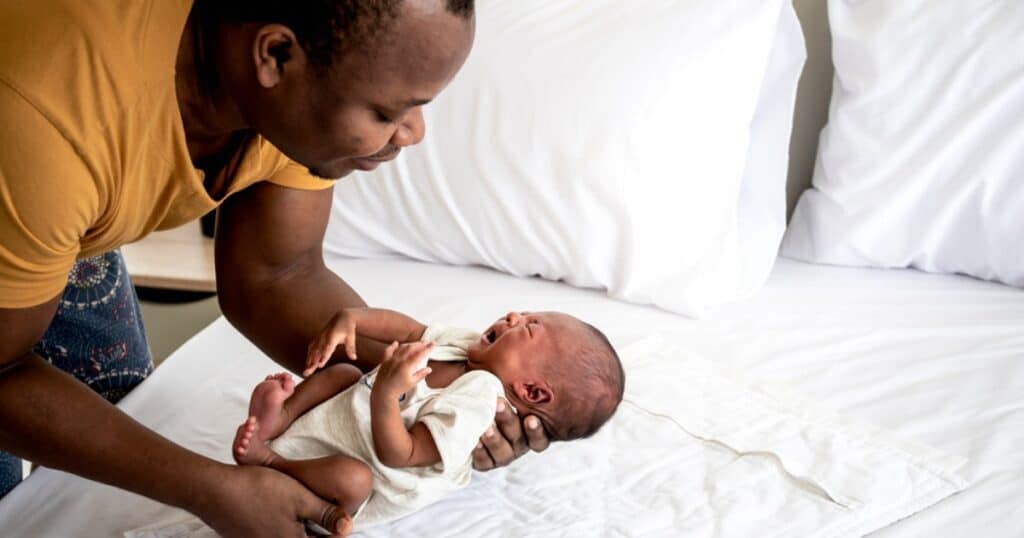 father holding crying infant next to bed